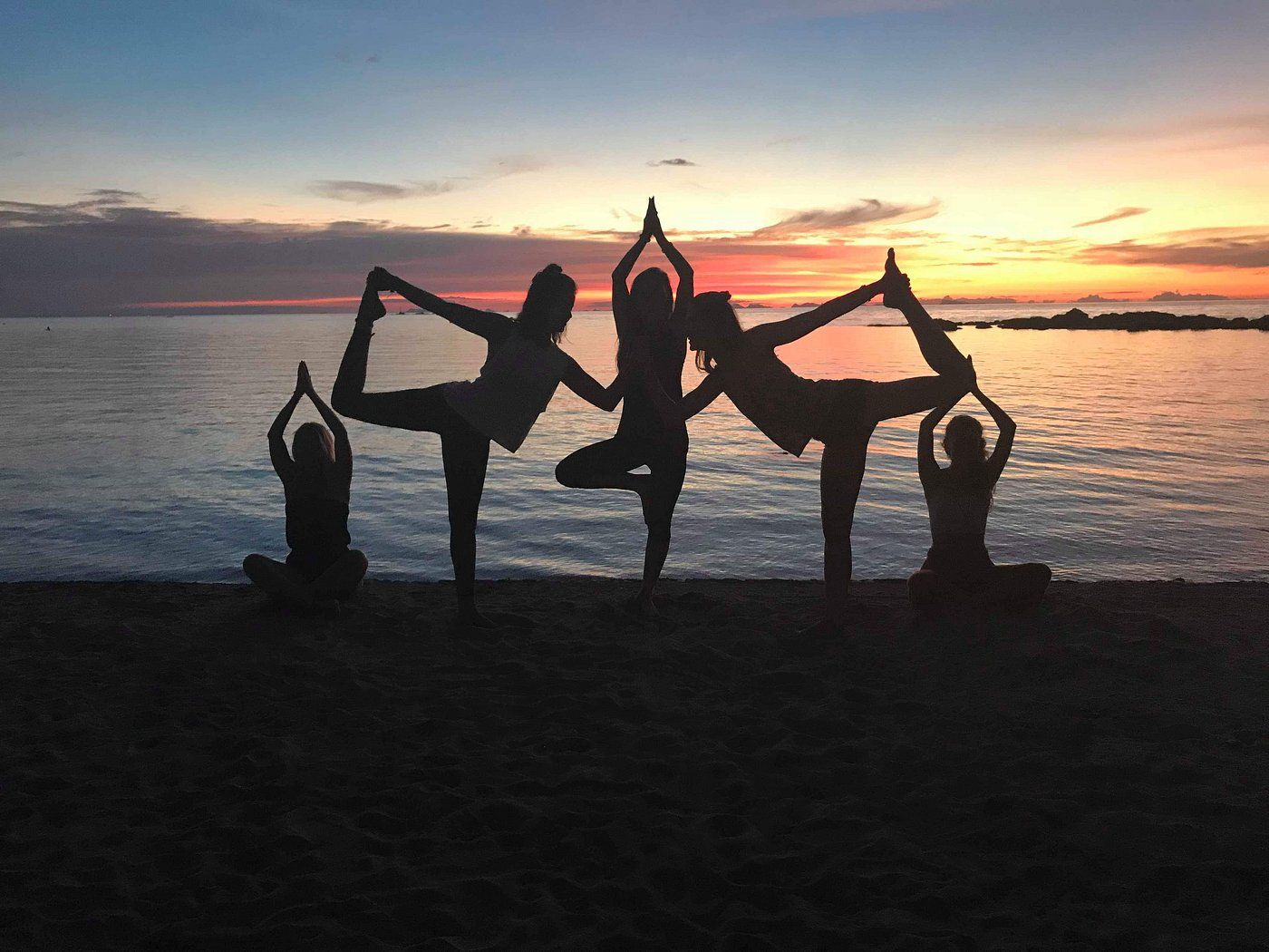 Four silhouetted people doing yoga poses at sunset by the water