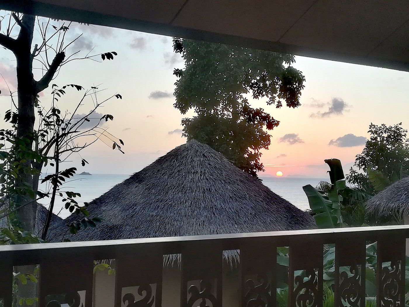 Sunset over the ocean behind a thatched roof and balcony, framed by silhouetted trees and plants