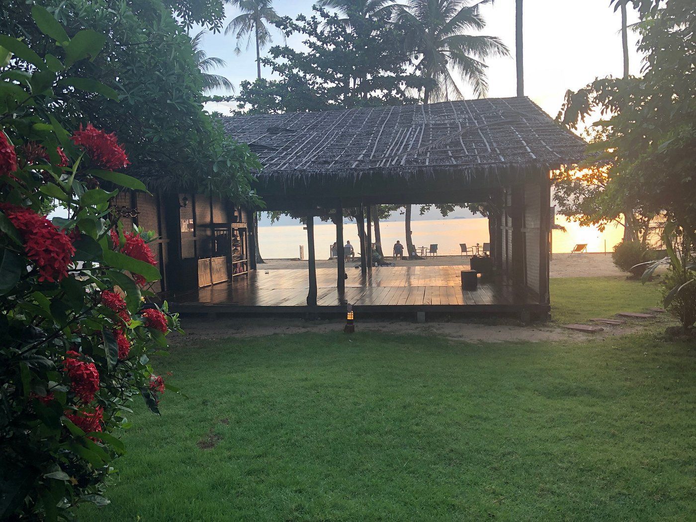 Sunset over a thatched pavilion by a grassy lawn, with palm trees and ocean in the background