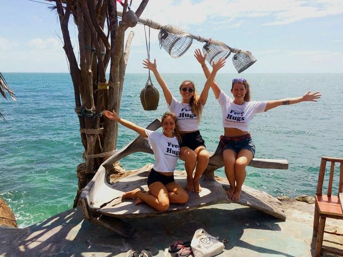 Three people posing on a seaside deck, smiling and raising their arms beside the ocean.