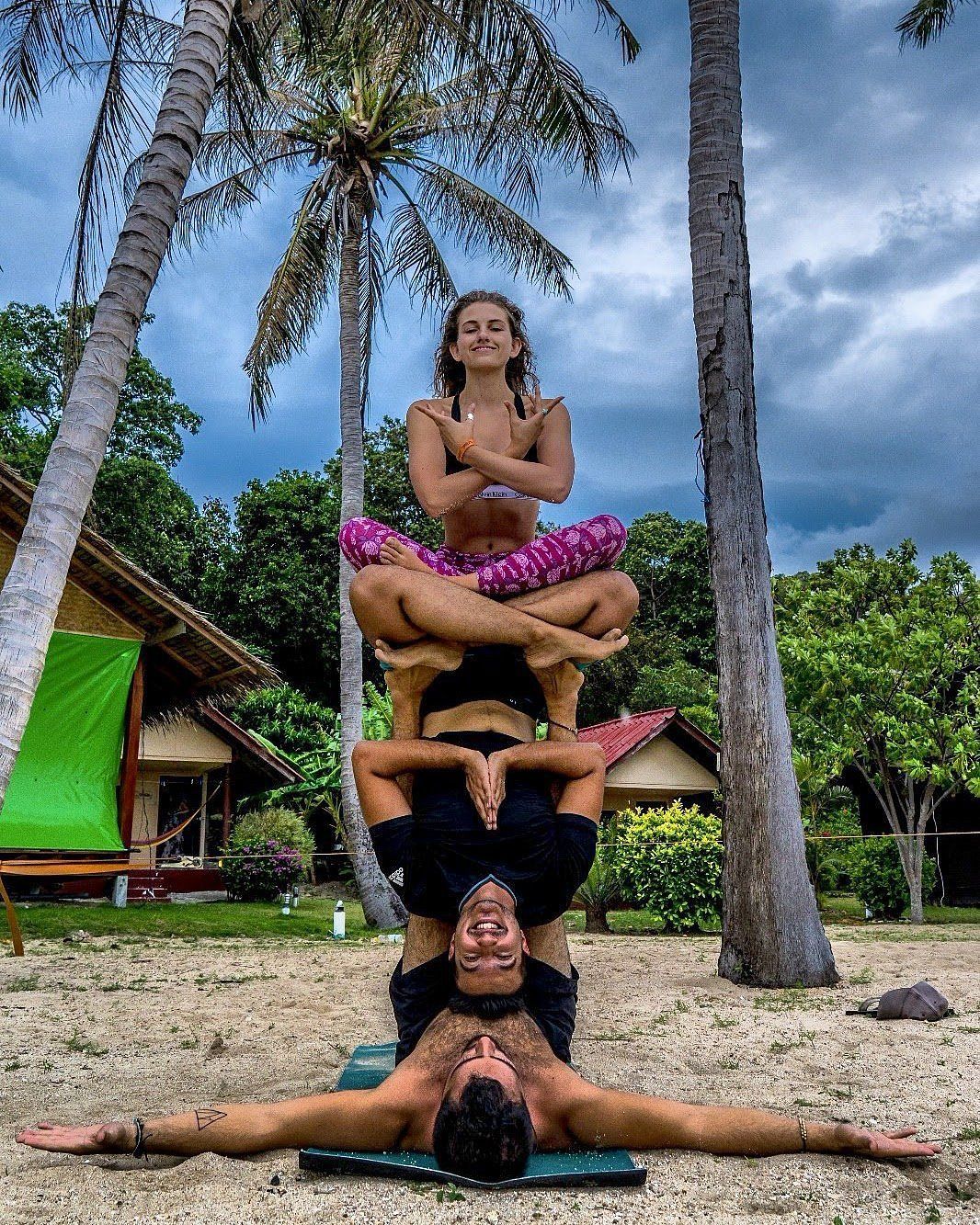 Four people balancing in a yoga pose on a beach, with palms and cloudy sky in the background.