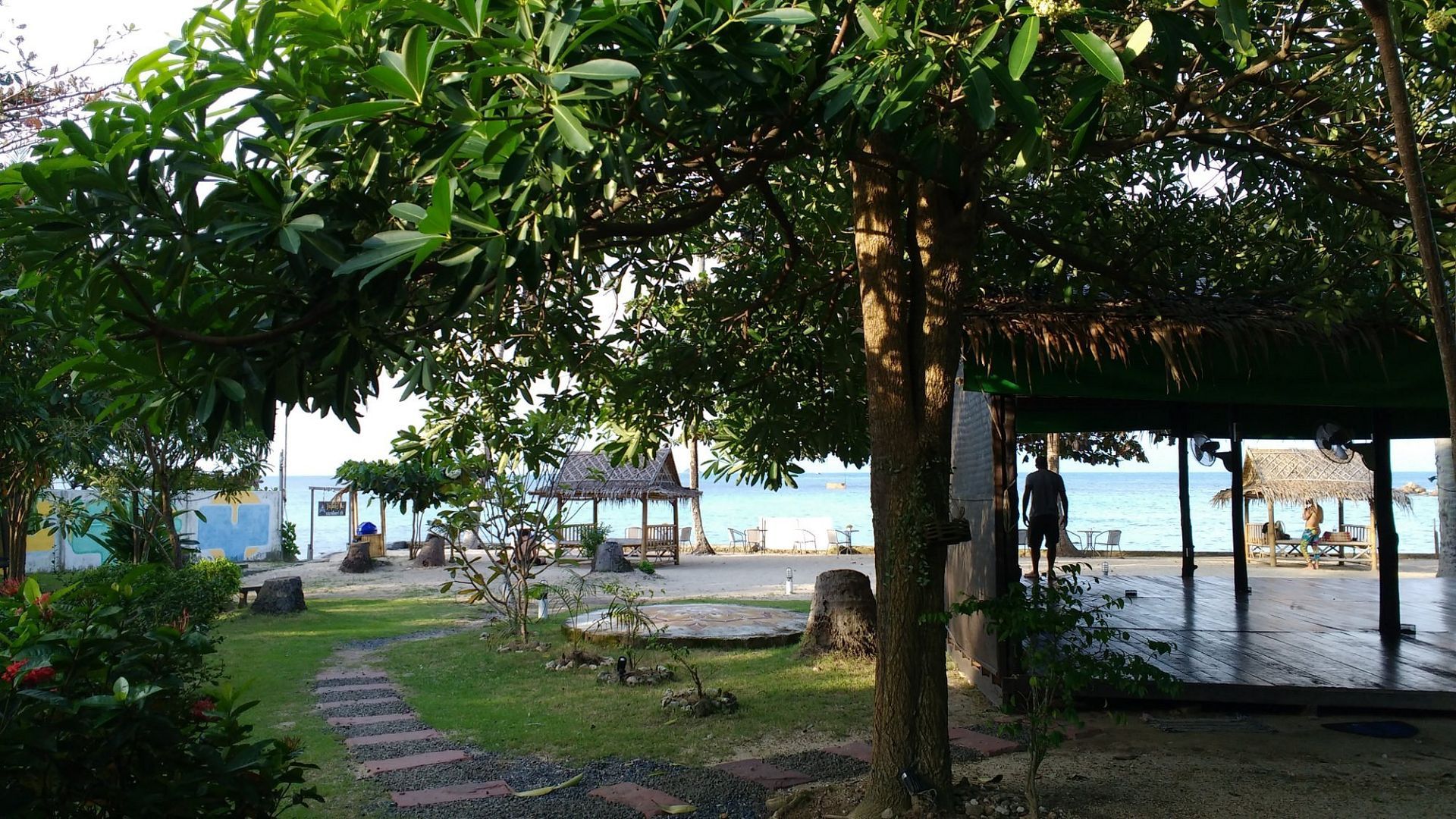 Shaded park with large trees, a stone path, and a small pavilion near the water at sunset