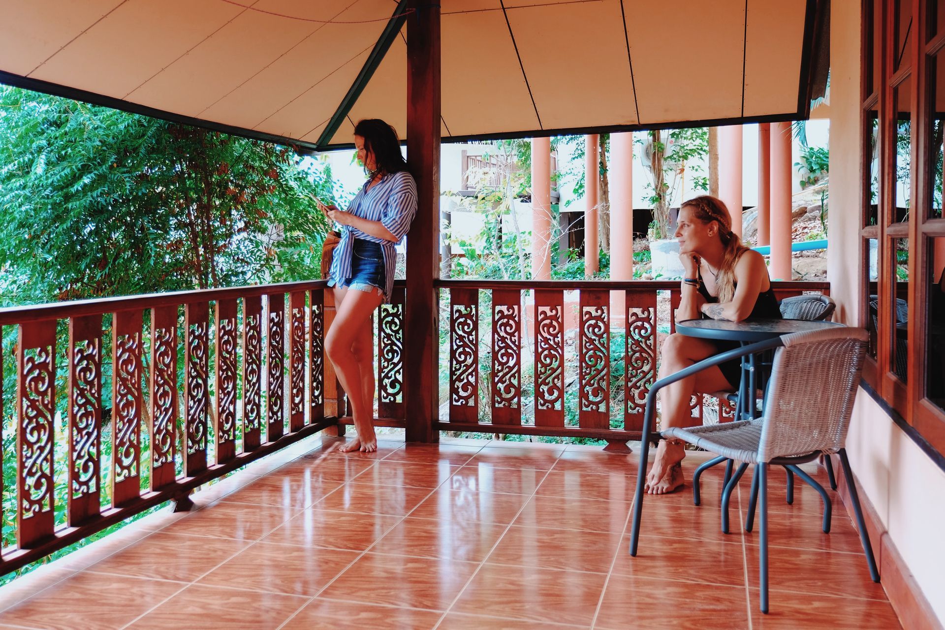 Two people on a wooden balcony, one leaning on the railing and one sitting at a table with chairs.
