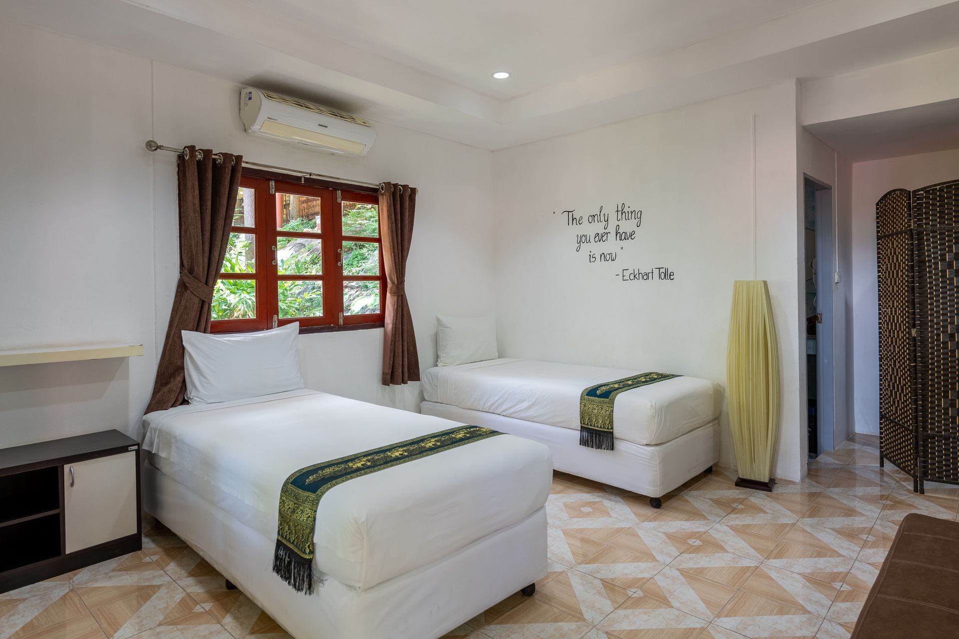 Bright bedroom with two twin beds, patterned tile floor, window, and wall-mounted air conditioner.
