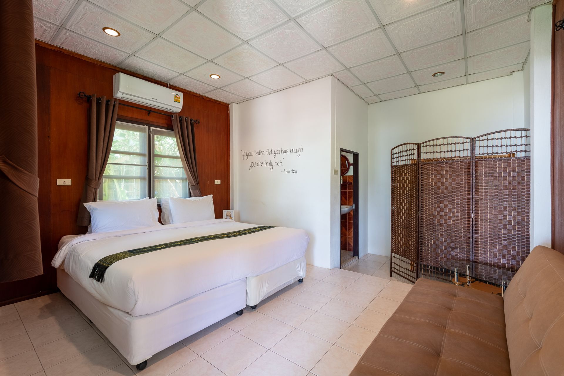 Bright hotel room with a white bed, window curtains, air conditioner, and wicker room divider