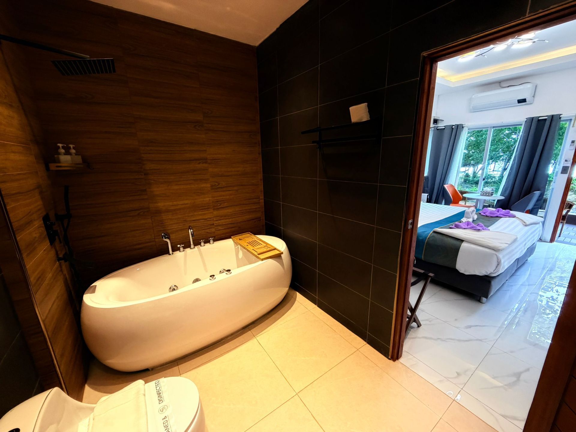 Modern bathroom with a freestanding tub and dark tiled walls, opening to a bright bedroom beyond.
