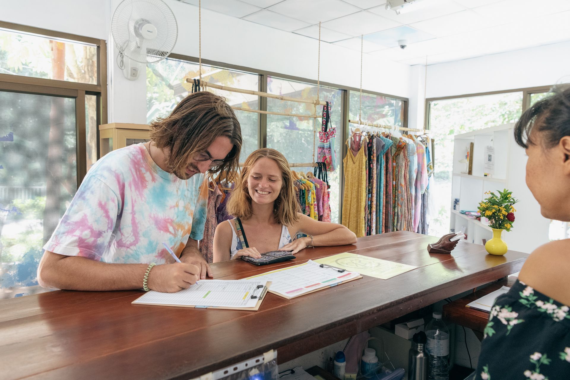 Two people signing papers at a shop counter, with clothing racks and a vase of flowers nearby.