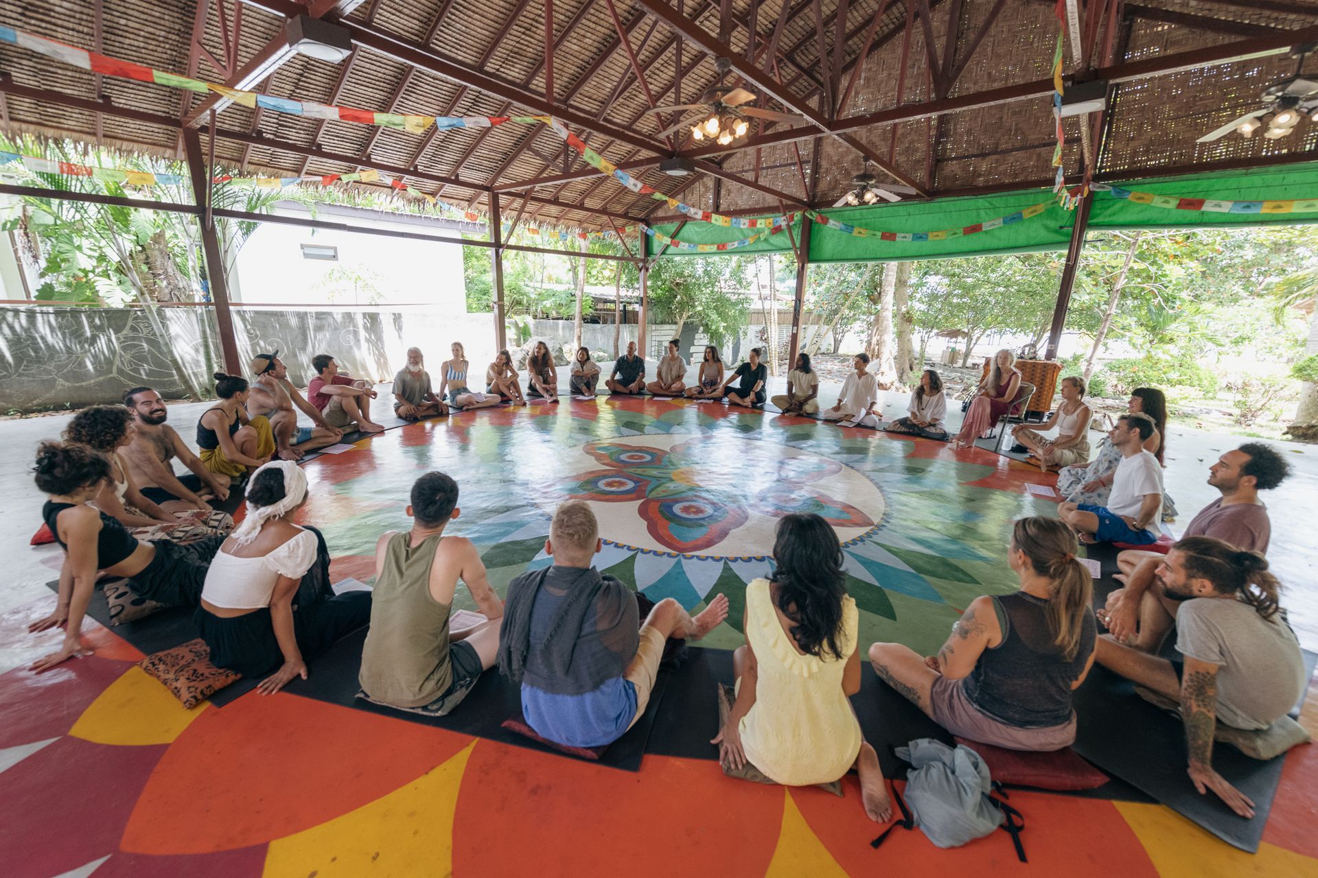 People seated in a circle on mats in an open-air pavilion, gathering around a central painted floor.