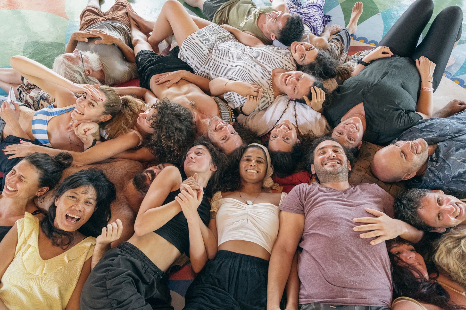 Group of smiling people lying in a circle on a wooden floor, hands reaching toward the center.