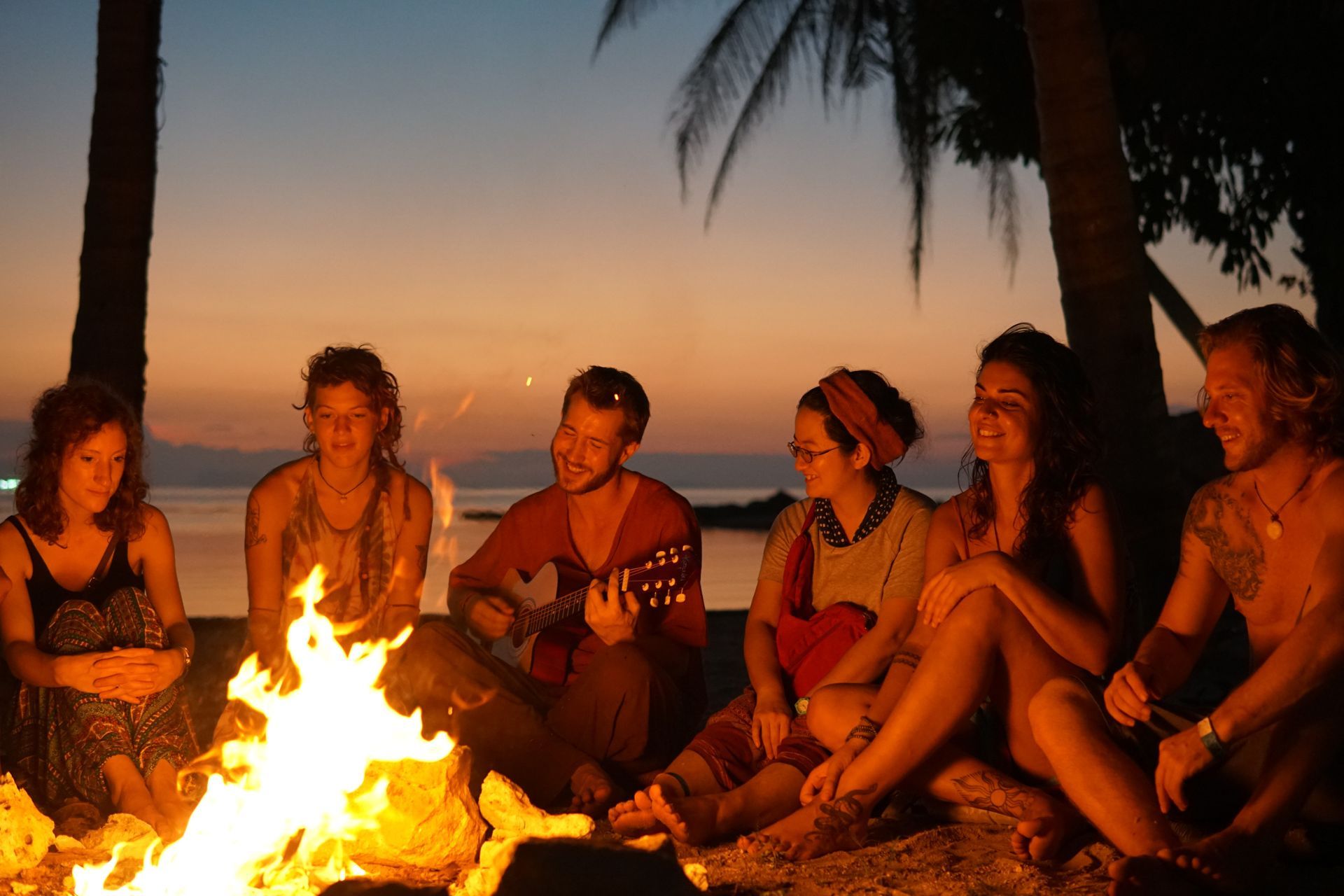 People sitting around a campfire at sunset on a beach, with palm trees in the background