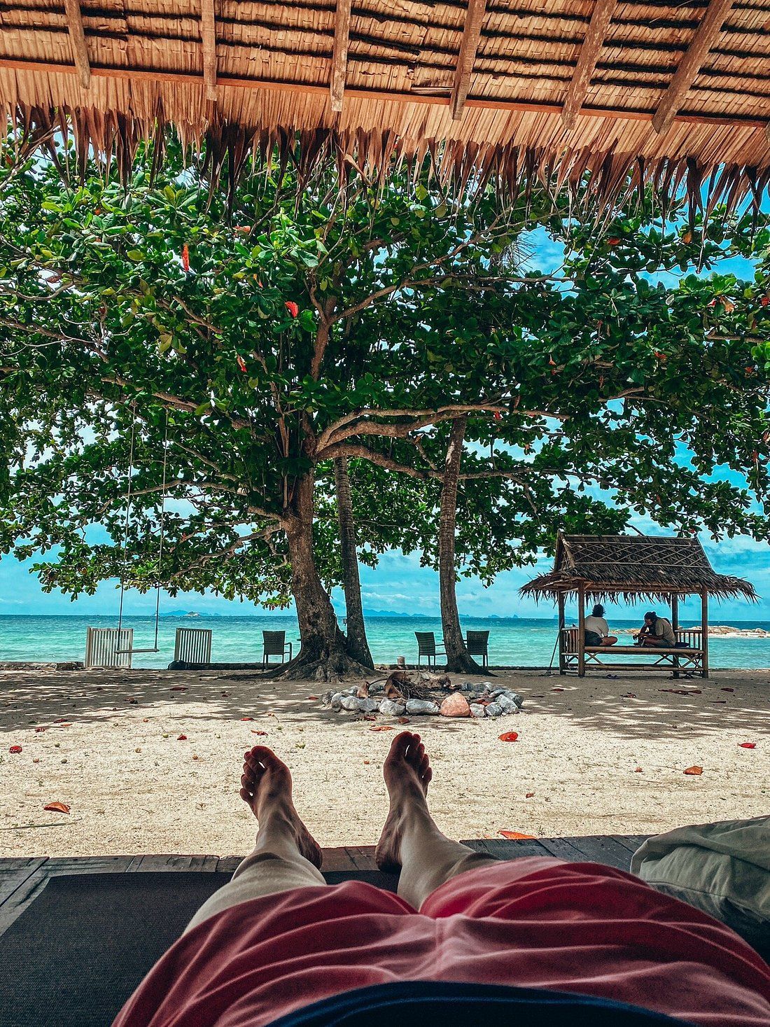 Relaxing beach view from a hammock, with feet visible, turquoise water, sandy shore, trees, and a thatched shelter.