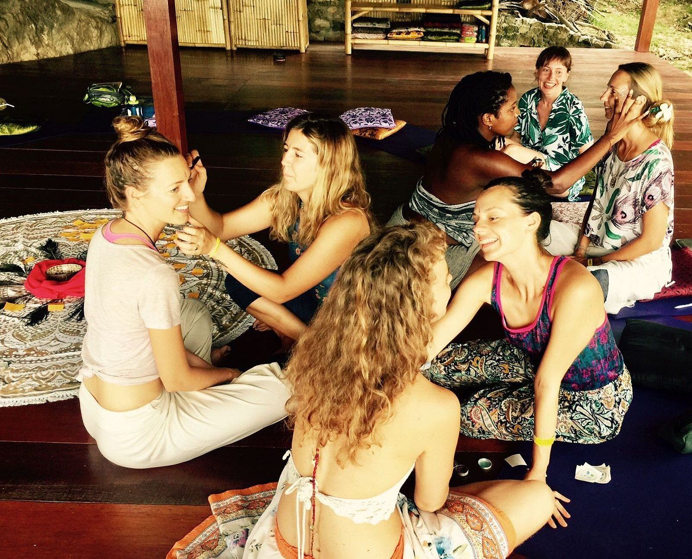 People sitting in a circle on woven mats, talking and smiling in a shaded outdoor space.