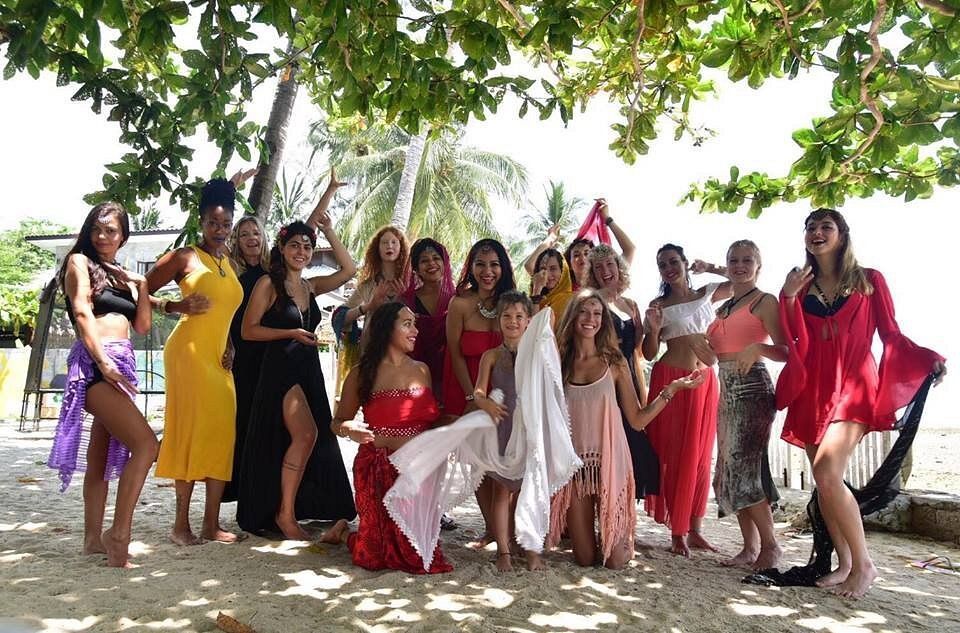 Group posing outdoors under trees, many in colorful dresses, with a fountain and beach in the background