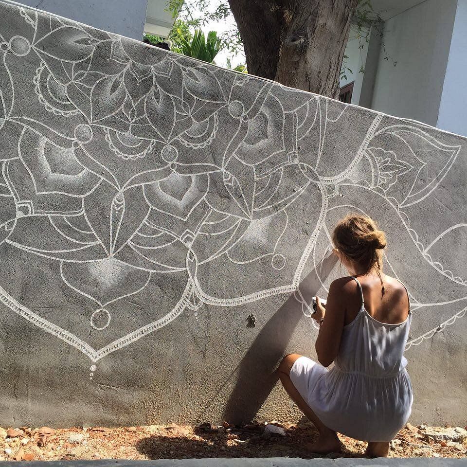 Person drawing white chalk floral patterns on a large gray wall outdoors.