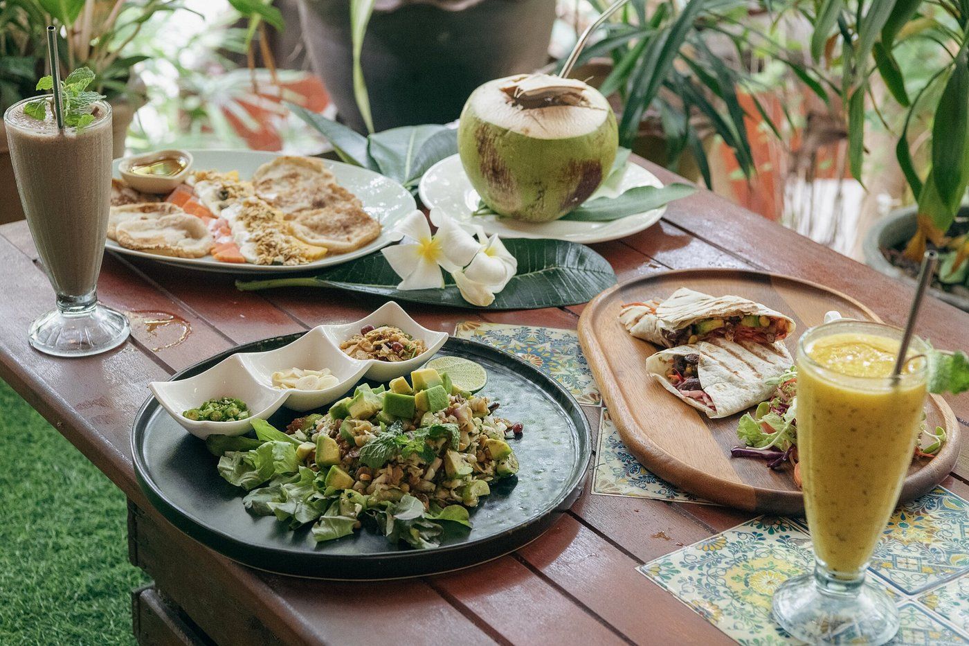 Plates of food and tropical drinks on a wooden table in a sunny outdoor café setting