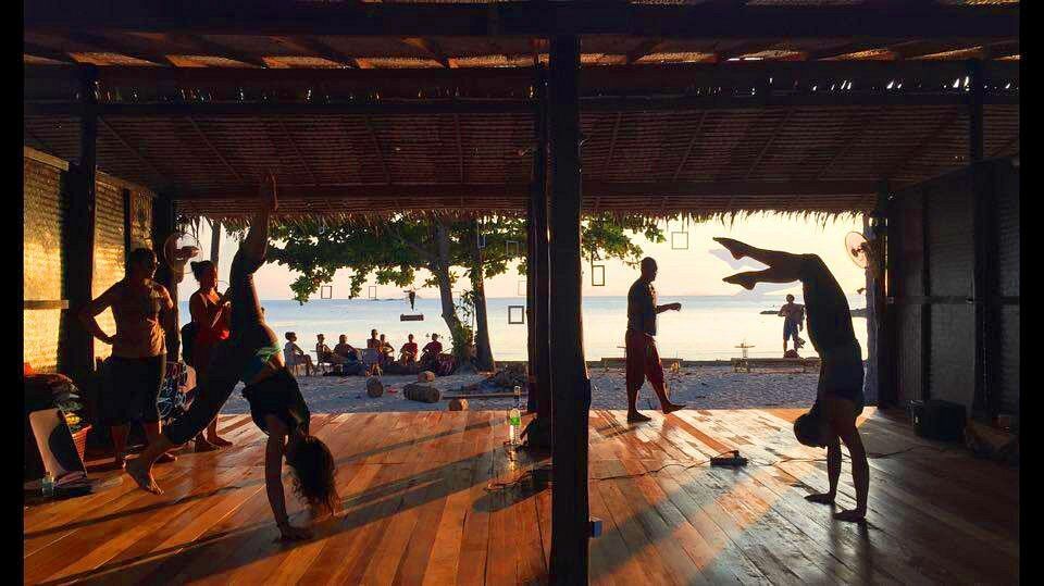 People practicing acrobatics in a wooden pavilion with a bright sunset and water view beyond.