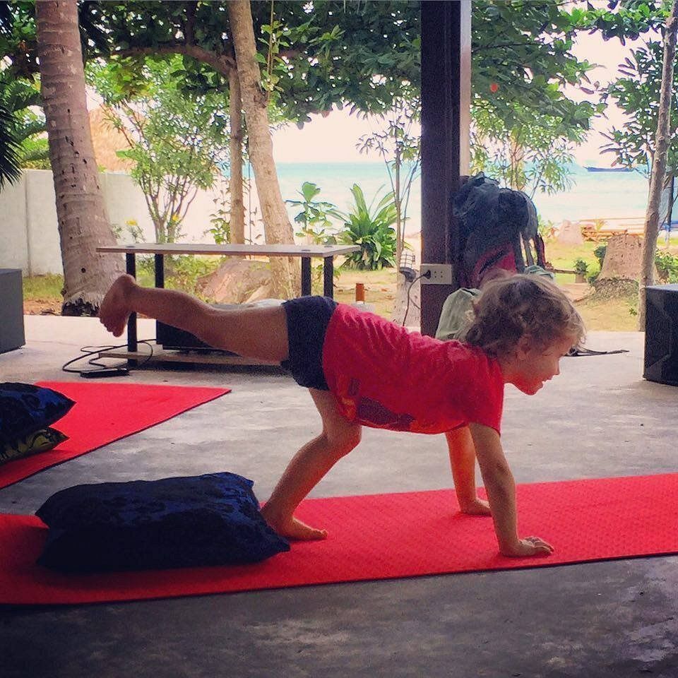 Person doing a one-arm balance pose on a red yoga mat in an open-air space by the beach