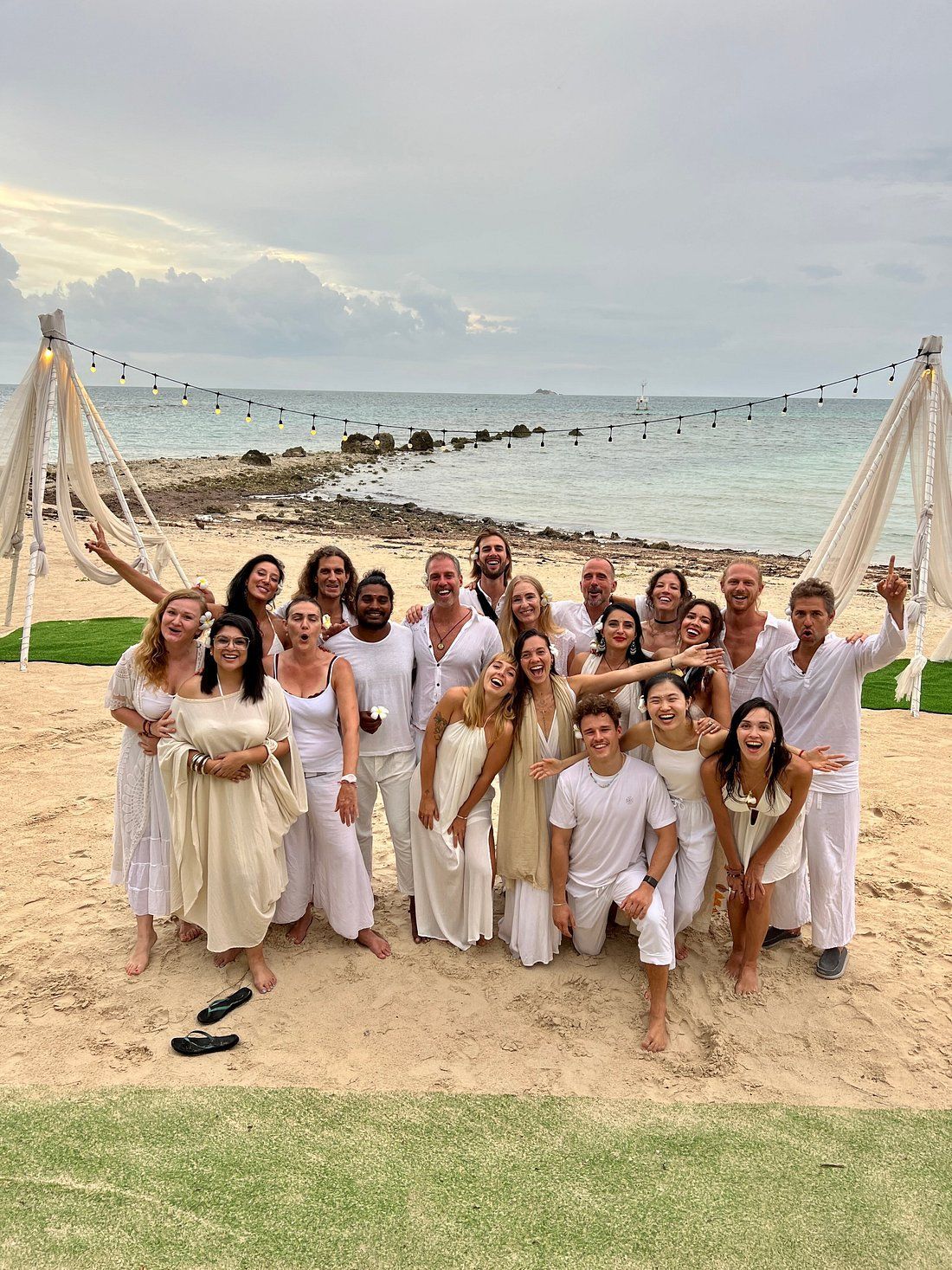 Group of people posing on a beach at sunset in white outfits, smiling near a decorated arch.