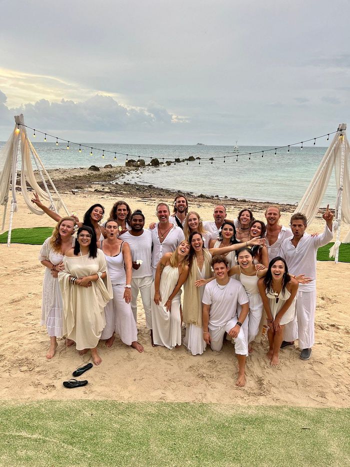 Group posing in white outfits at a beach wedding arch by the ocean, smiling at sunset.