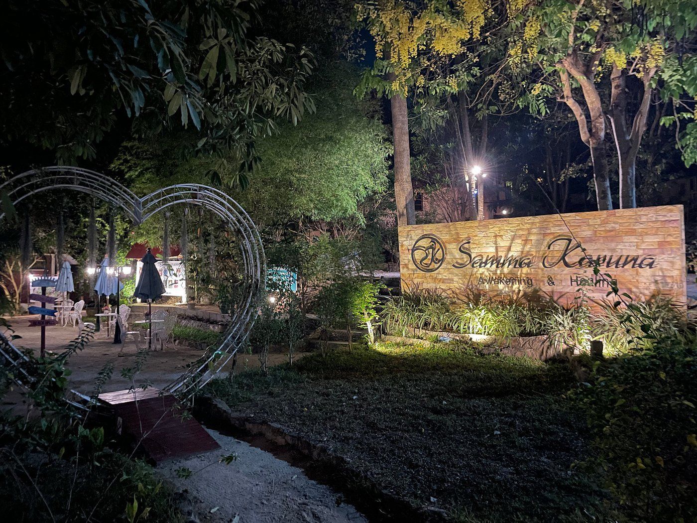 Nighttime garden entrance with lit archway, trees, and a wooden sign beside a path