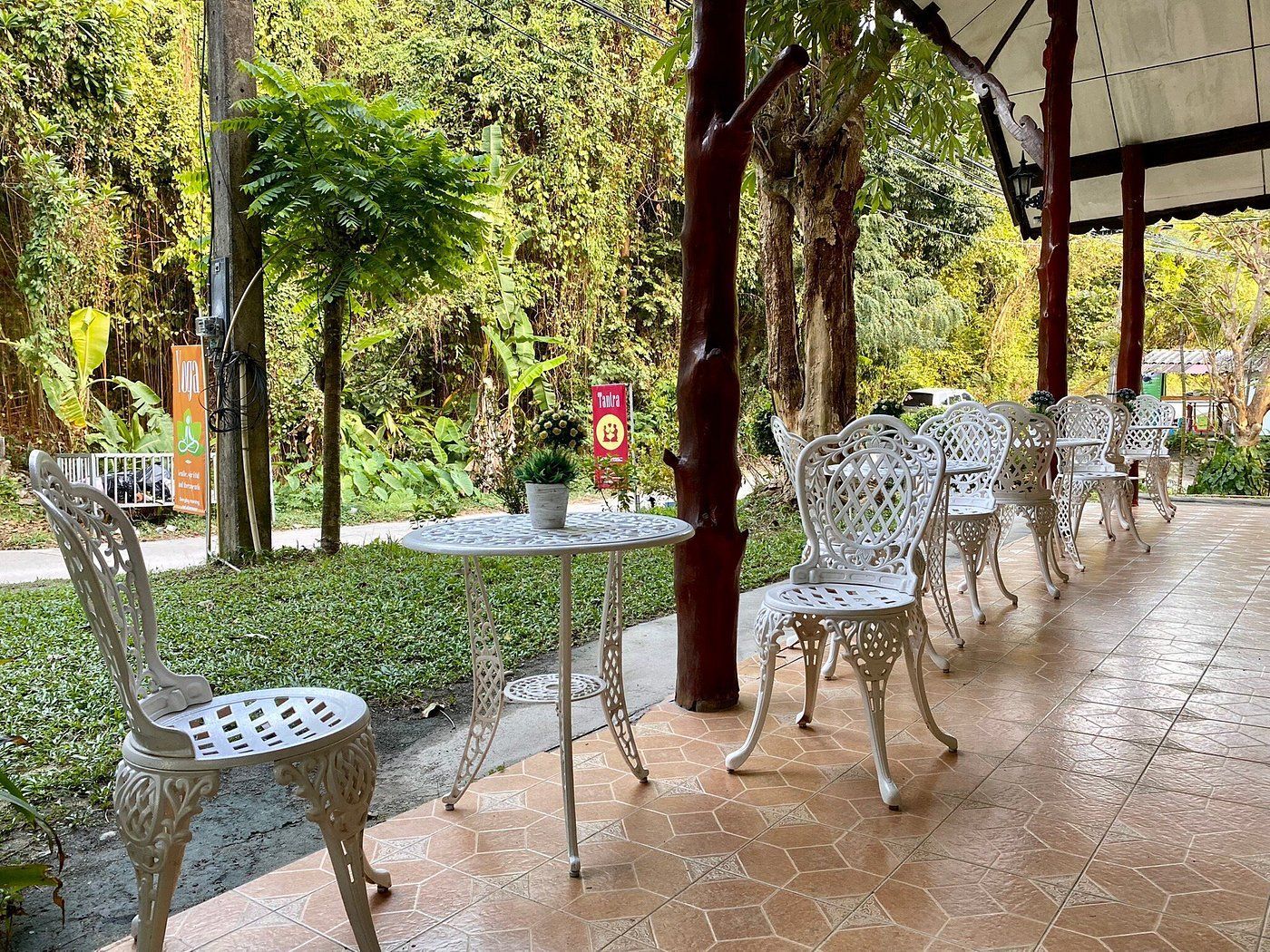 Outdoor café patio with white metal tables and chairs under trees, overlooking a lush garden.