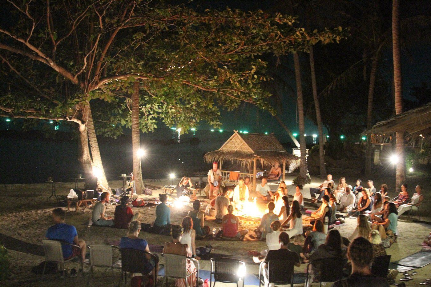 Nighttime outdoor gathering around a bonfire, with people seated under trees and string lights.