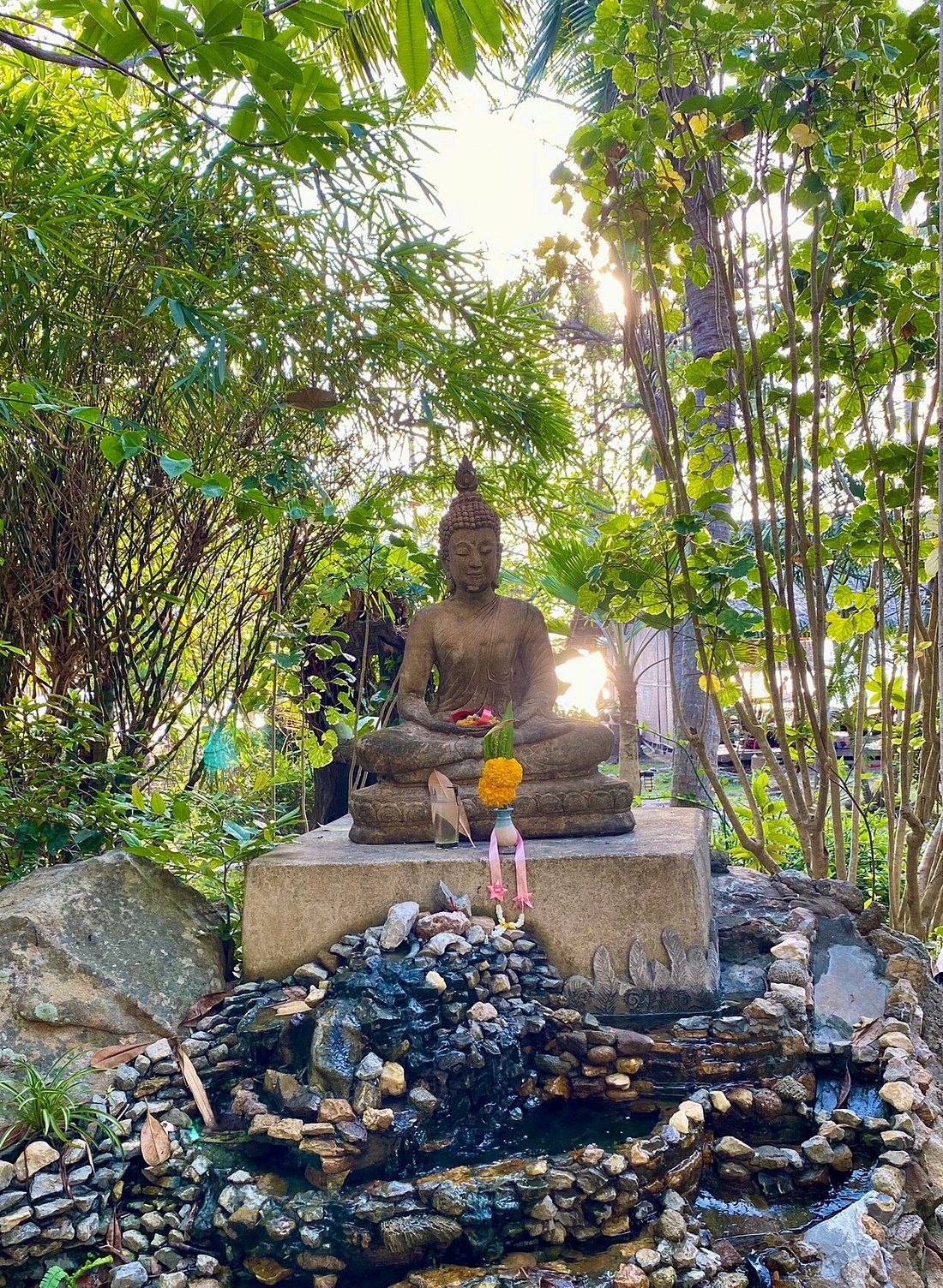 Buddha statue in a lush garden with incense and a small fire at the base