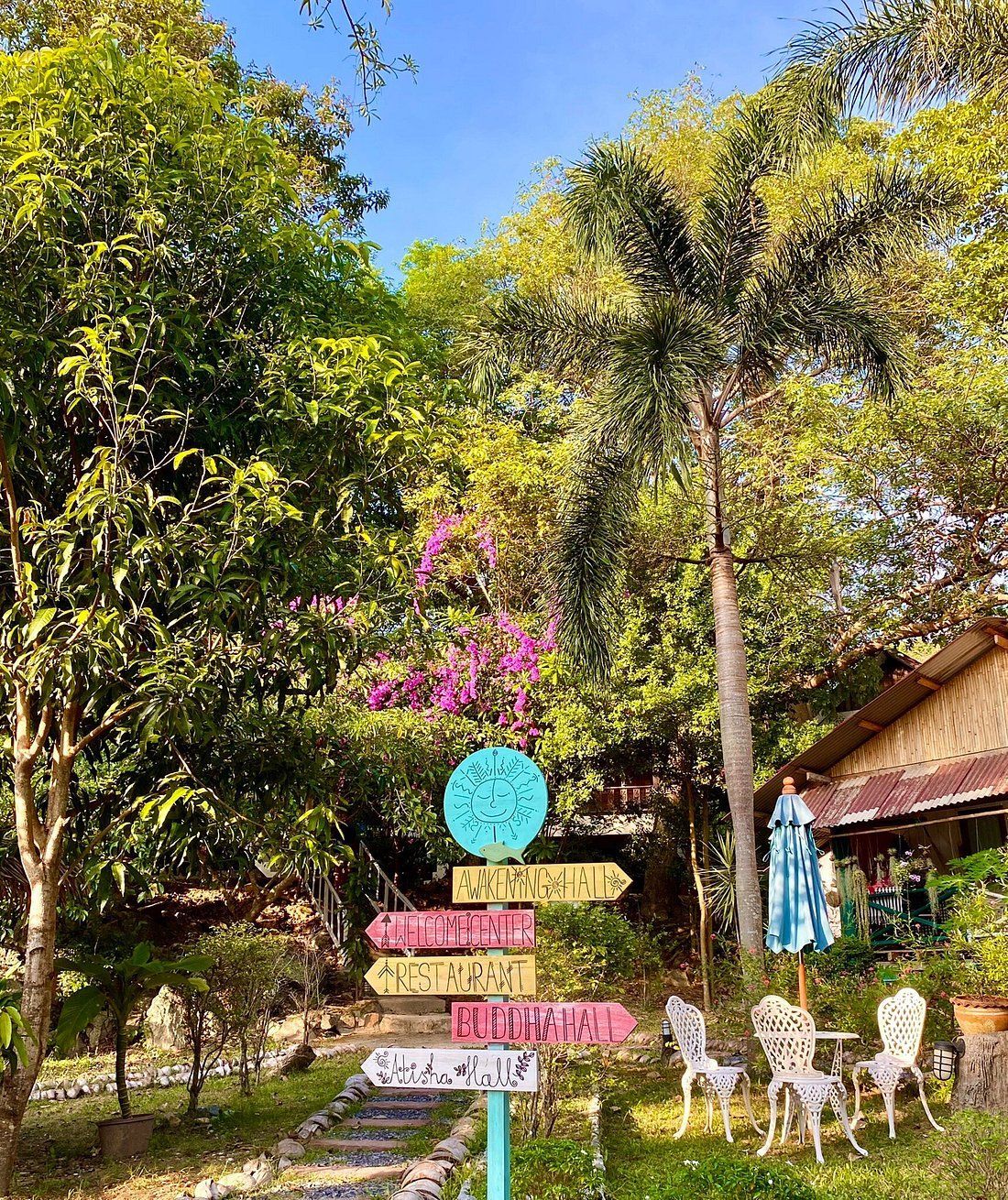Garden sign among trees and flowers beside a small patio with white chairs and a house