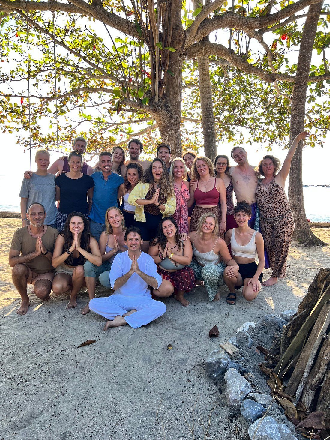 Group posing under a large tree on a beach, with sand, rocks, and a bright sunny setting.