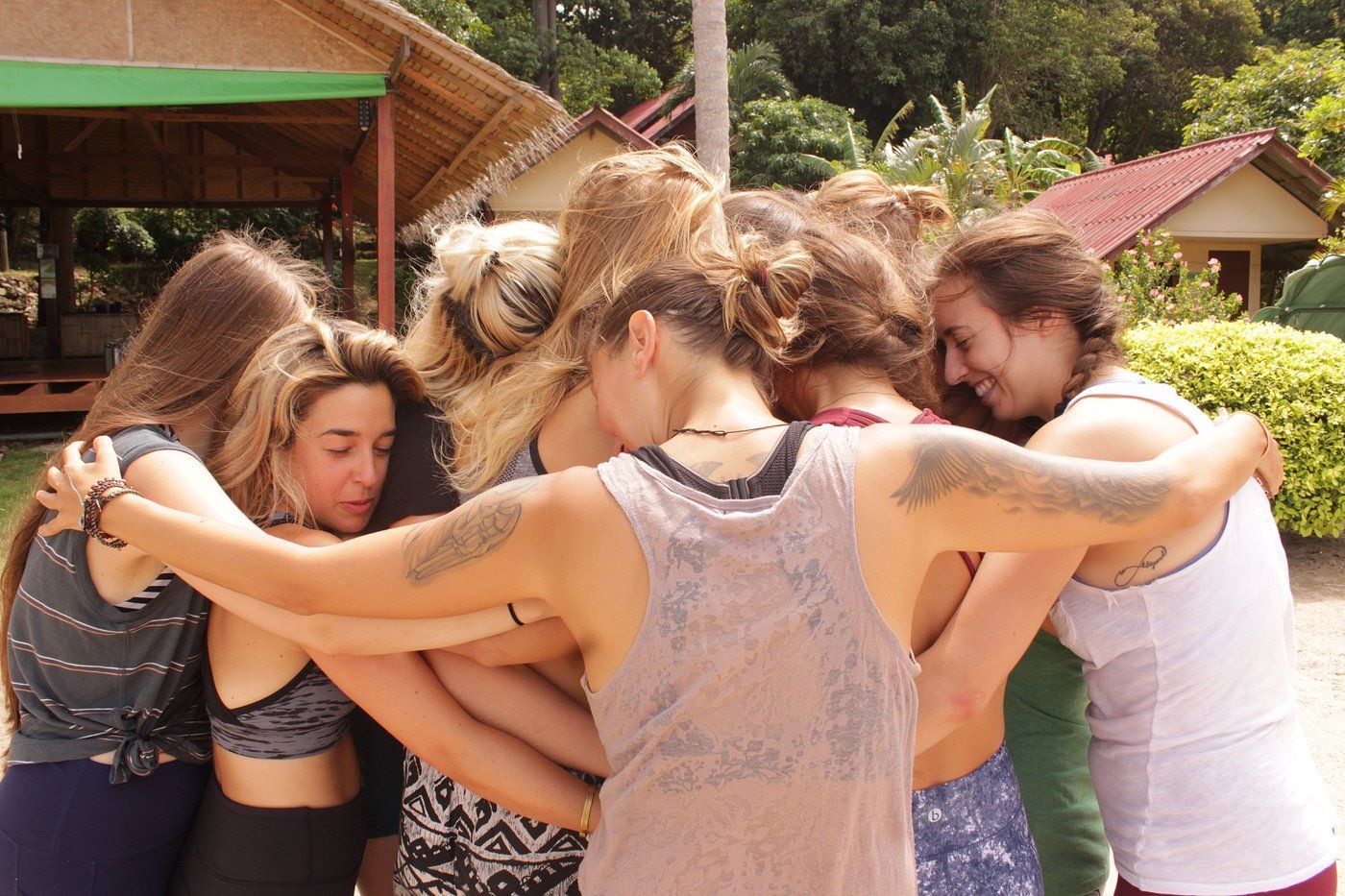 Group of people hugging in a circle outdoors near rustic buildings and trees
