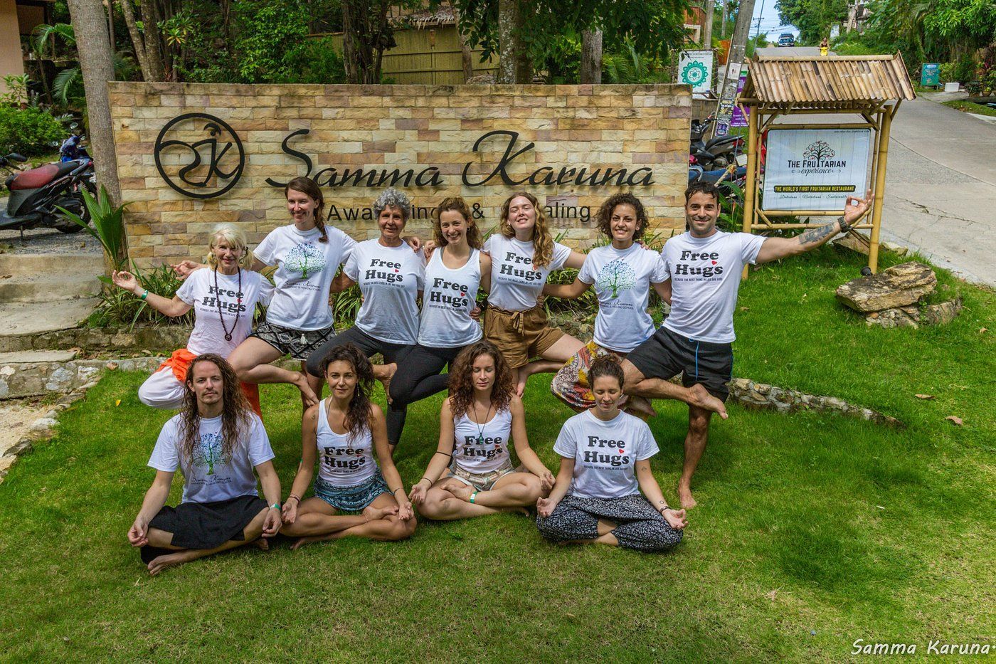 Group of people posing on a lawn in front of a sign, with a stone building and greenery behind them.