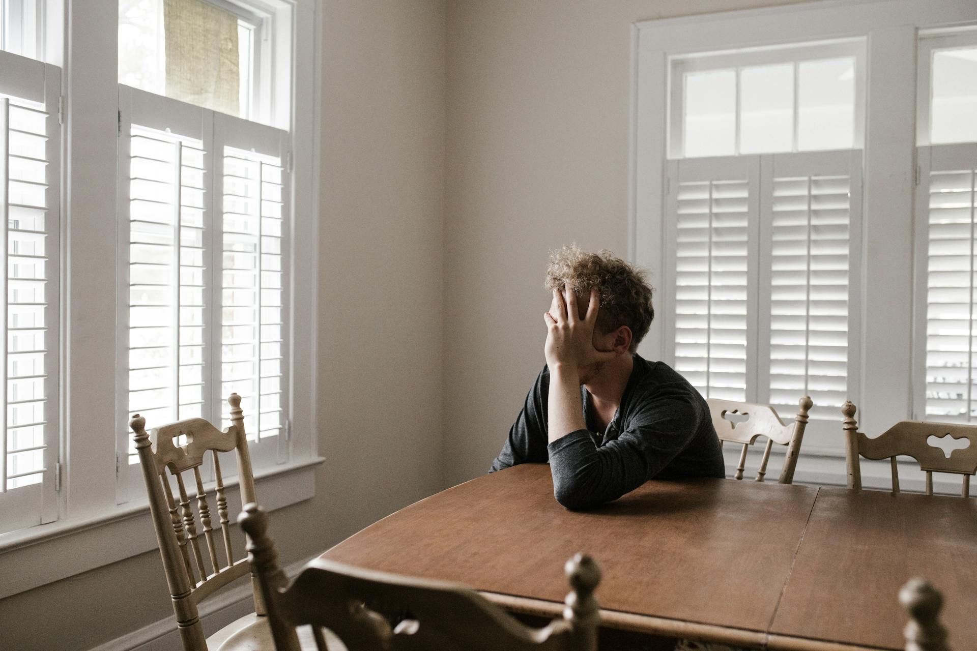 Anxiety Person at wooden table, head in hands, indoors, near windows with shutters.