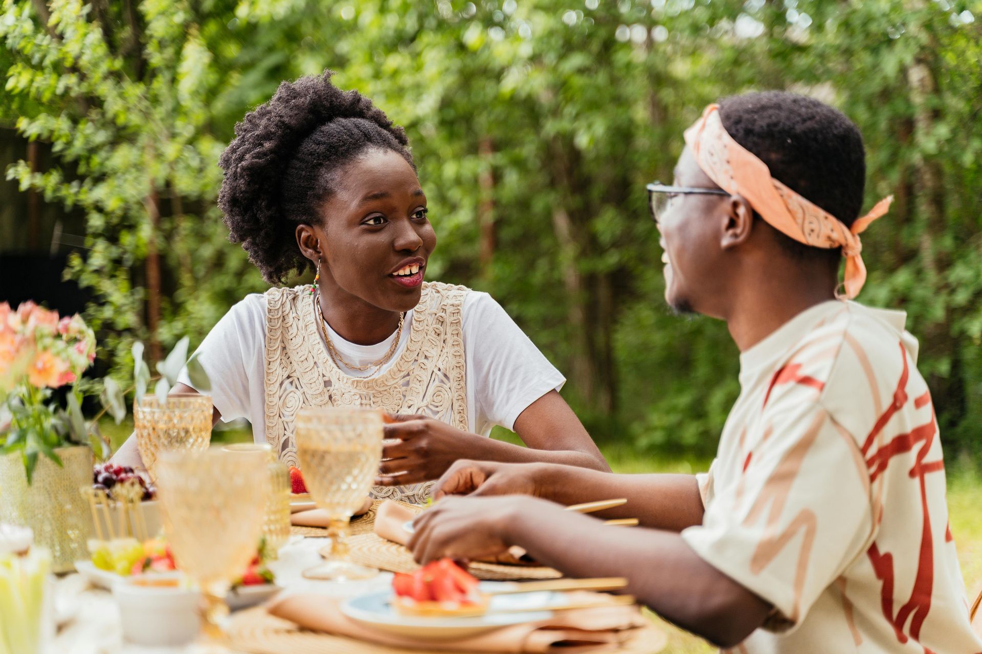 Two people having an outdoor picnic, smiling and talking while seated at a table with plates of food and drinks.