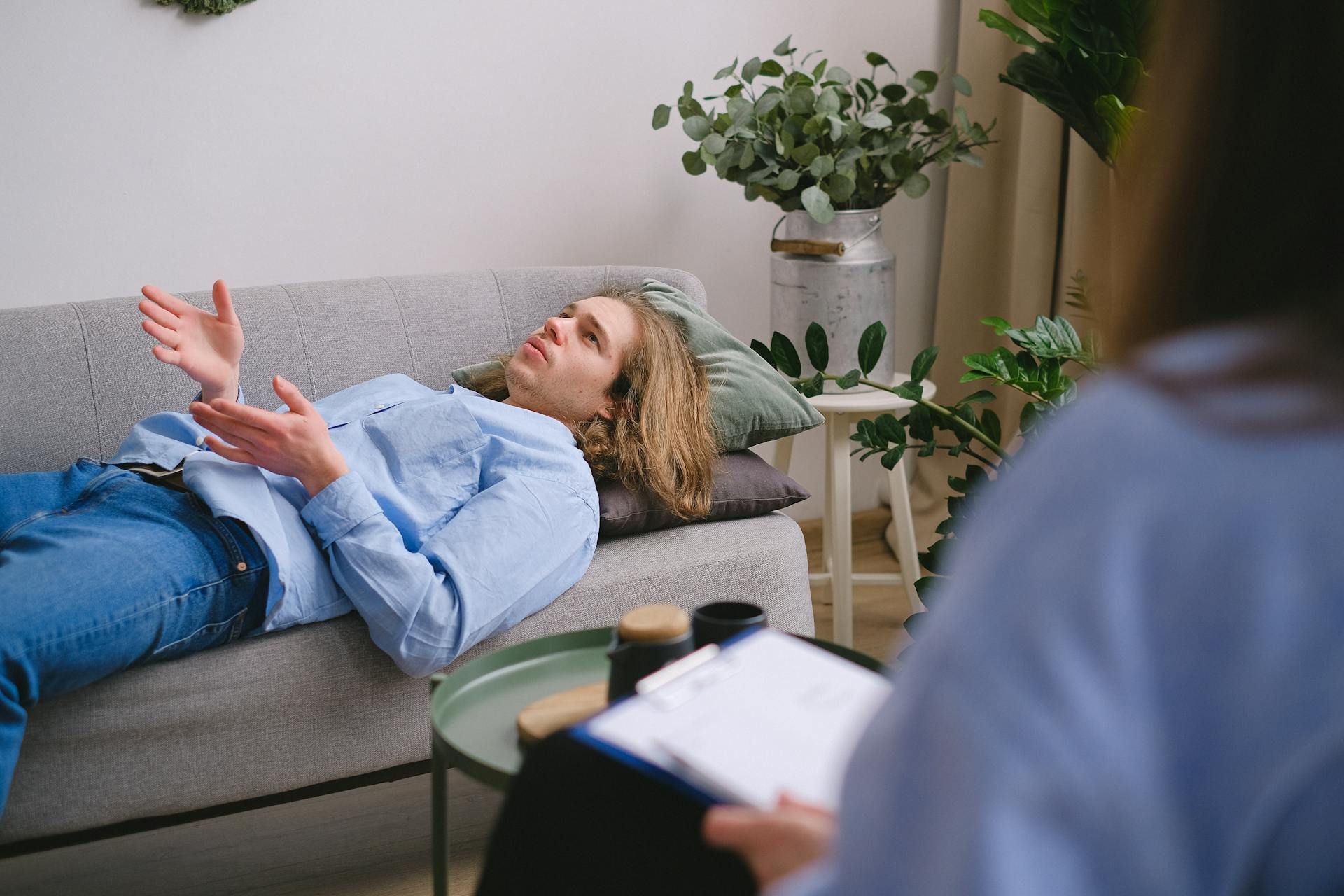 Man lying on a couch, talking to a therapist holding a clipboard. Indoor setting with plants.