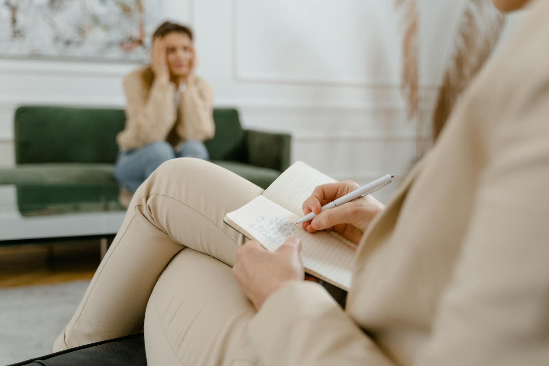 Therapist takes notes while patient clutches face, seated on green sofa.