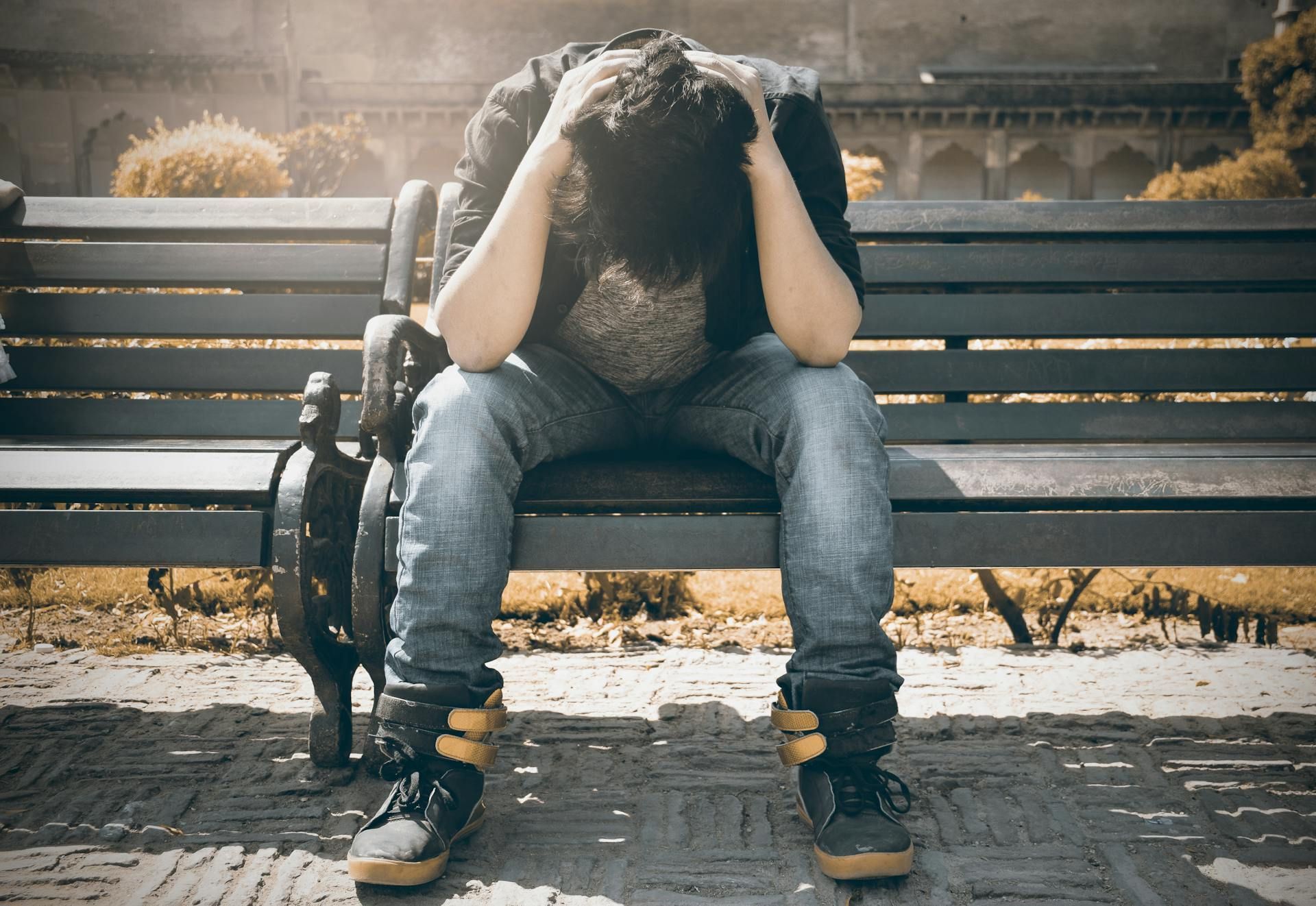 Person with head in hands, sitting on park bench, appearing distressed.