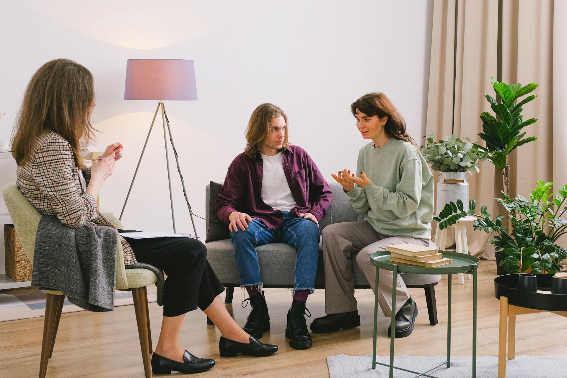 Three people in a counseling session; a person takes notes while two others sit and talk on a sofa with plants in the background.