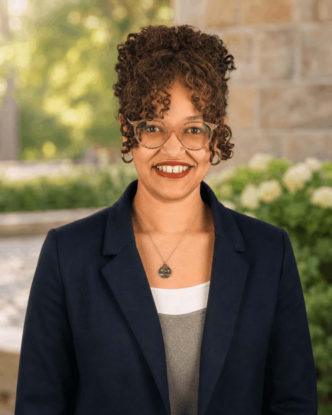 Portrait of a smiling person with curly hair and glasses, standing outdoors in a navy blazer
