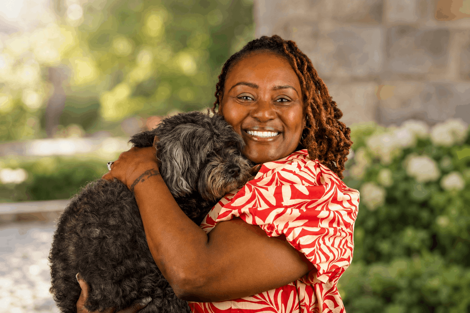 Smiling person hugging a small black dog outdoors with blurred greenery and flowers behind them