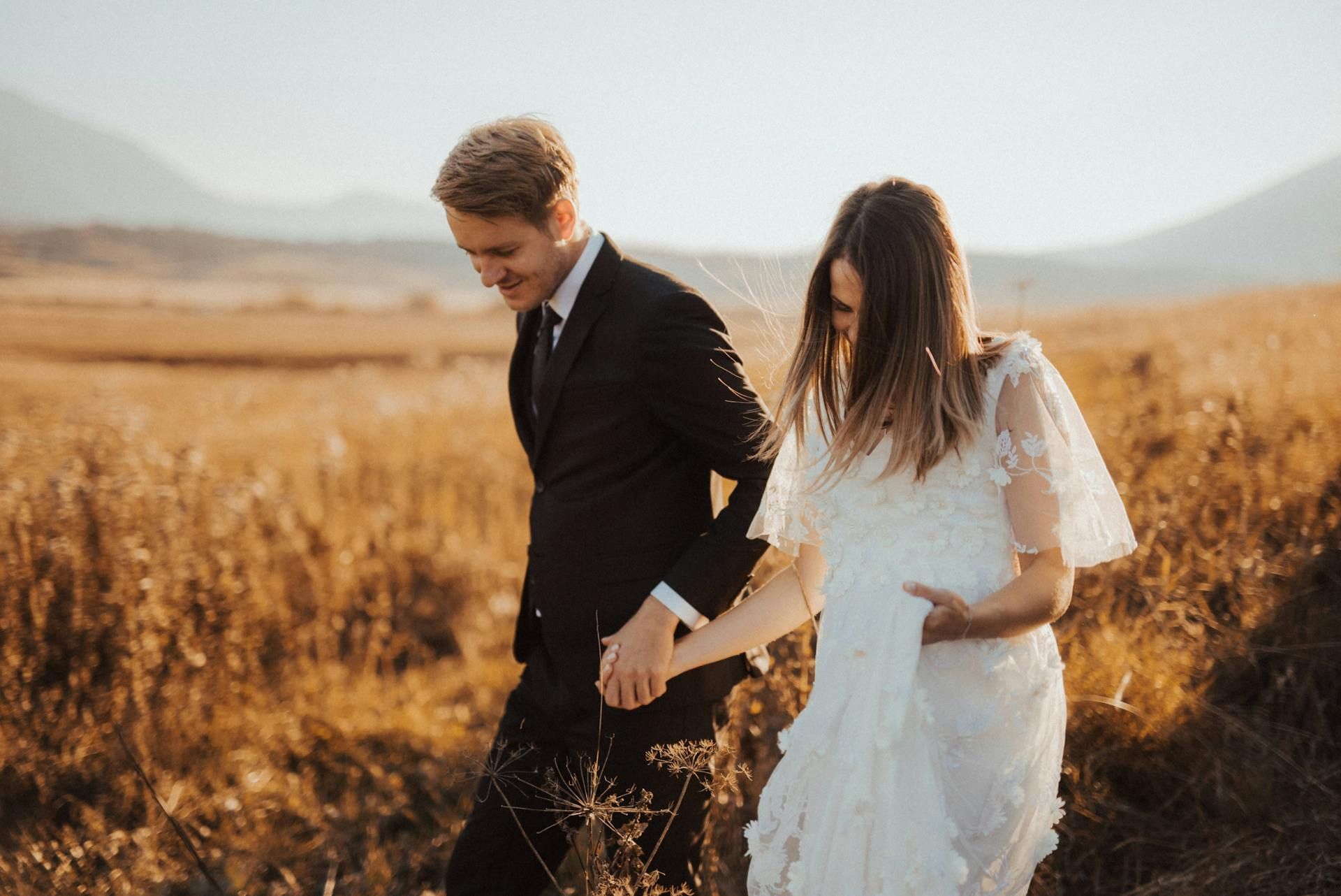 A couple in wedding attire walks hand-in-hand through a grassy, sunlit field during the golden hour.