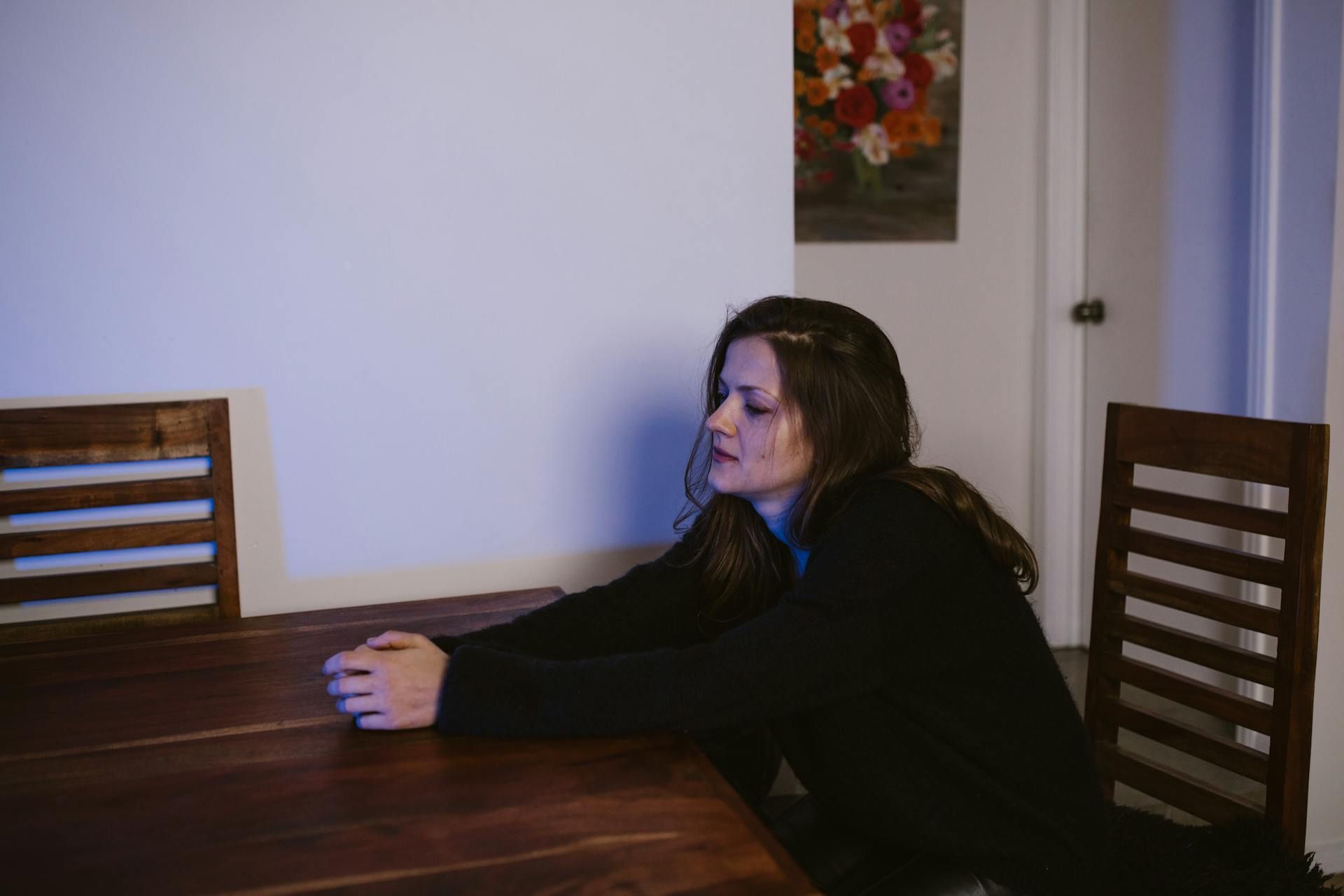 Woman seated at a wooden table, resting arms on it. She looks thoughtful. A painting and door are in the background.
