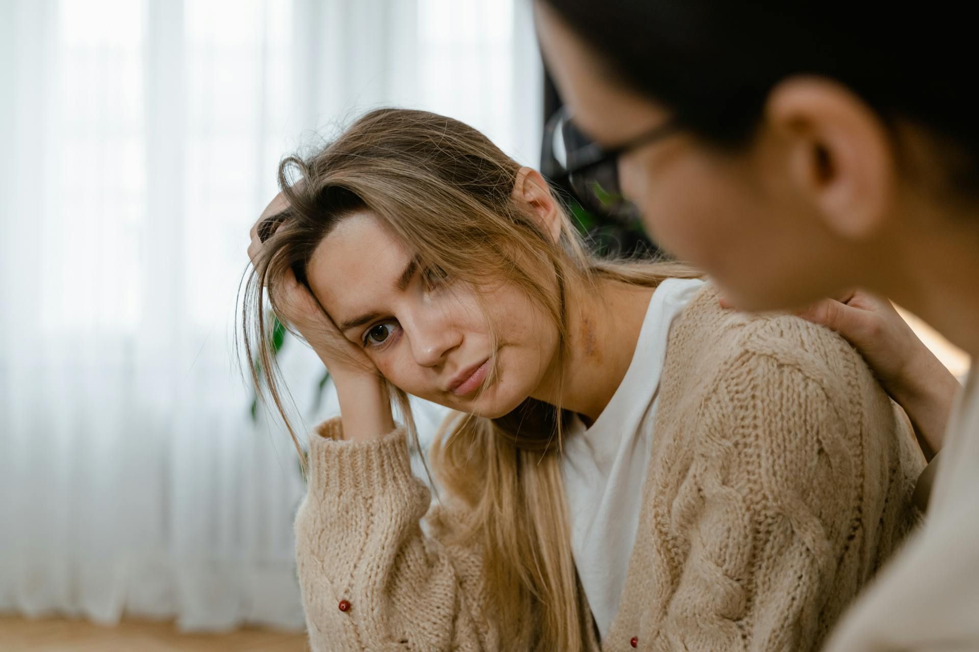 Woman in beige sweater looks sad, comforted by another person's hand on her shoulder.