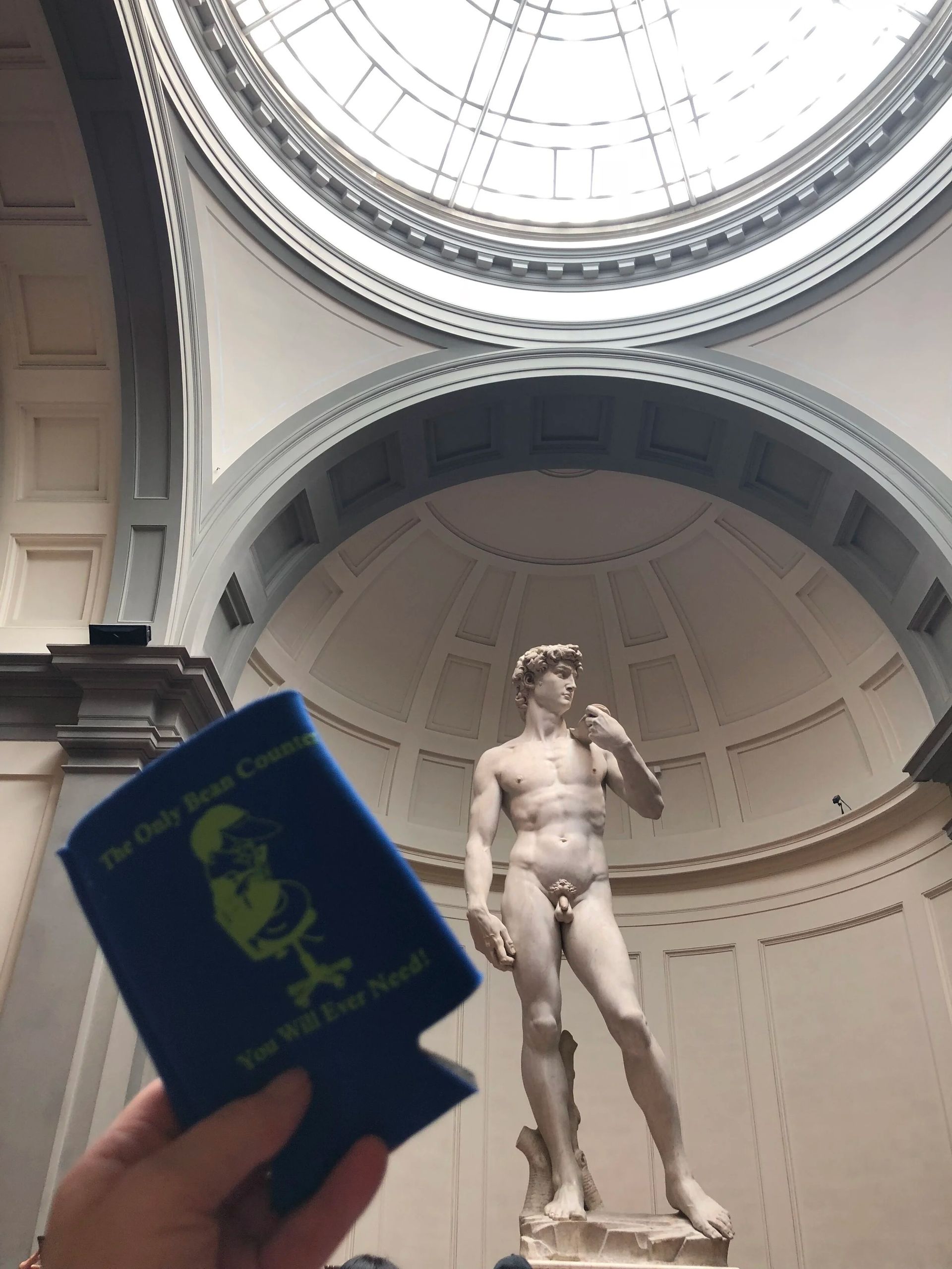Person holds blue drink sleeve in front of Michelangelo's David statue, inside a domed gallery.