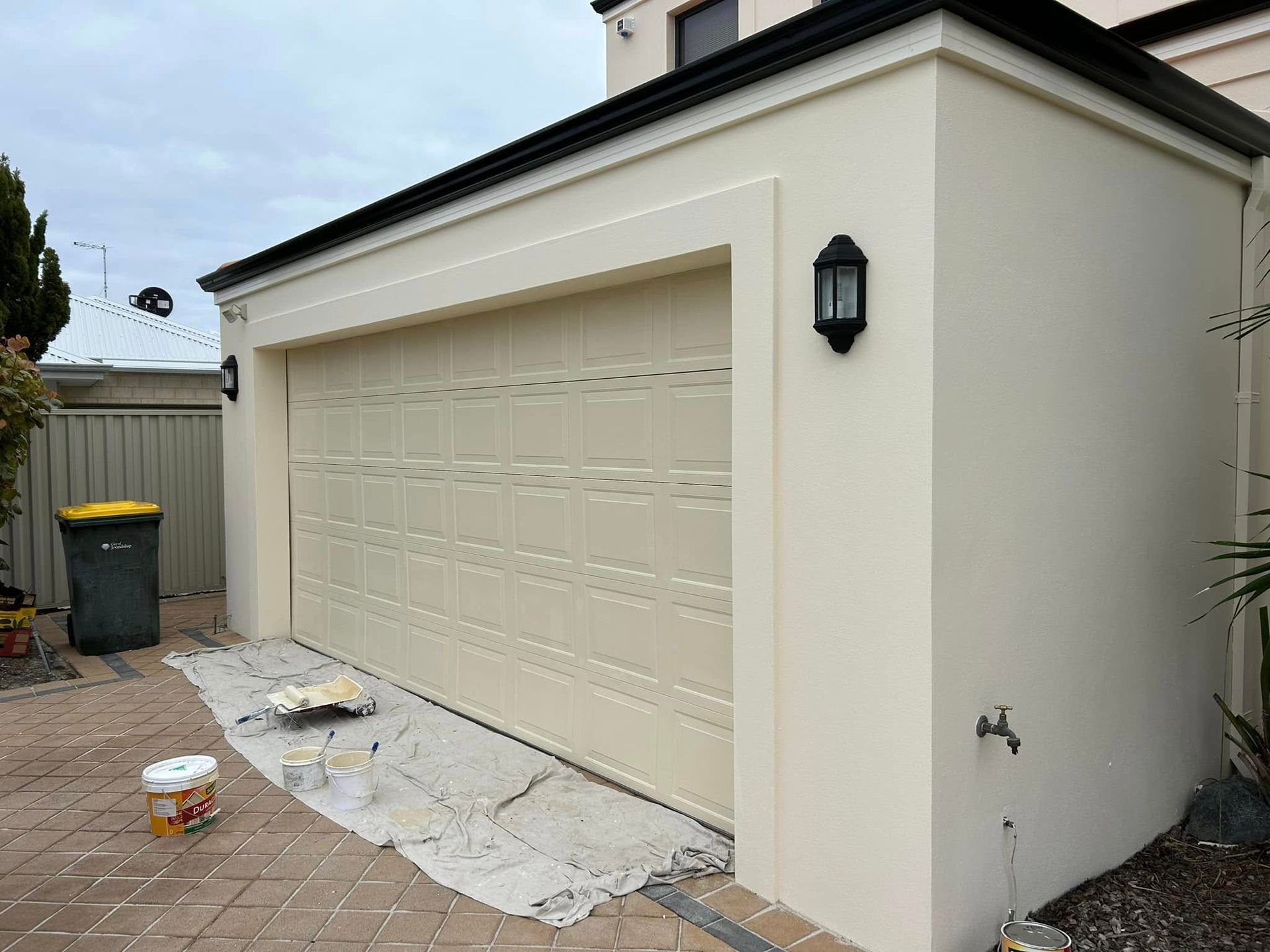 Cream garage door on a beige house, with paint supplies and a drop cloth on the driveway.