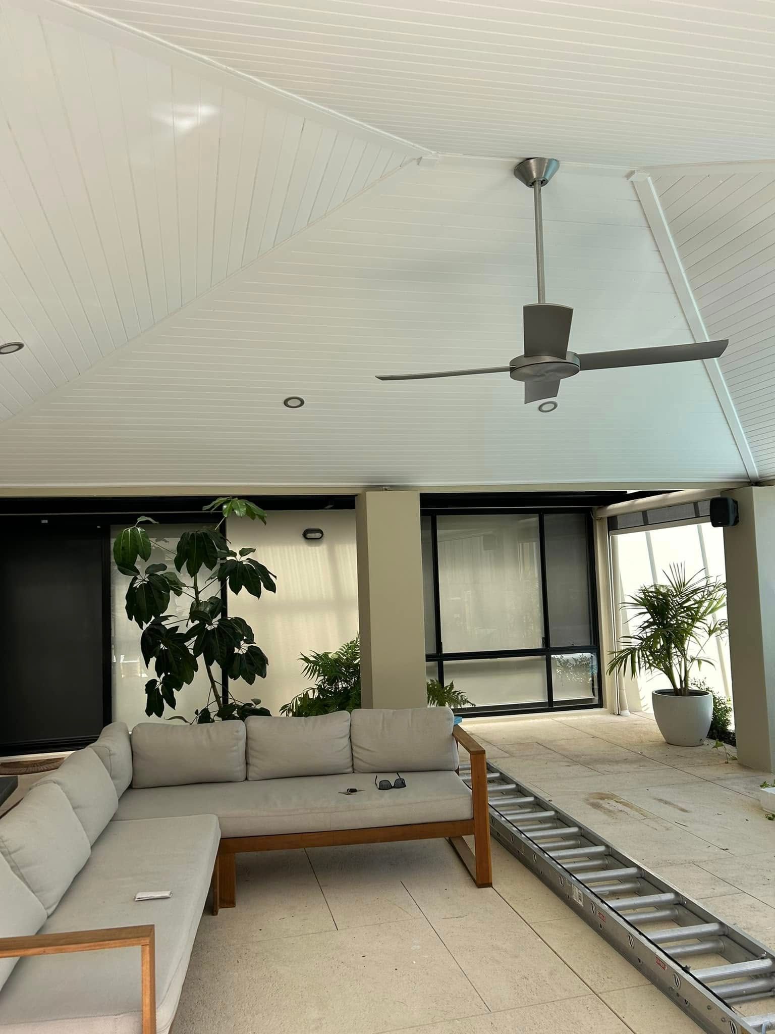Modern living room with L-shaped sofa, ceiling fan, large windows, plants, and a ladder on the floor