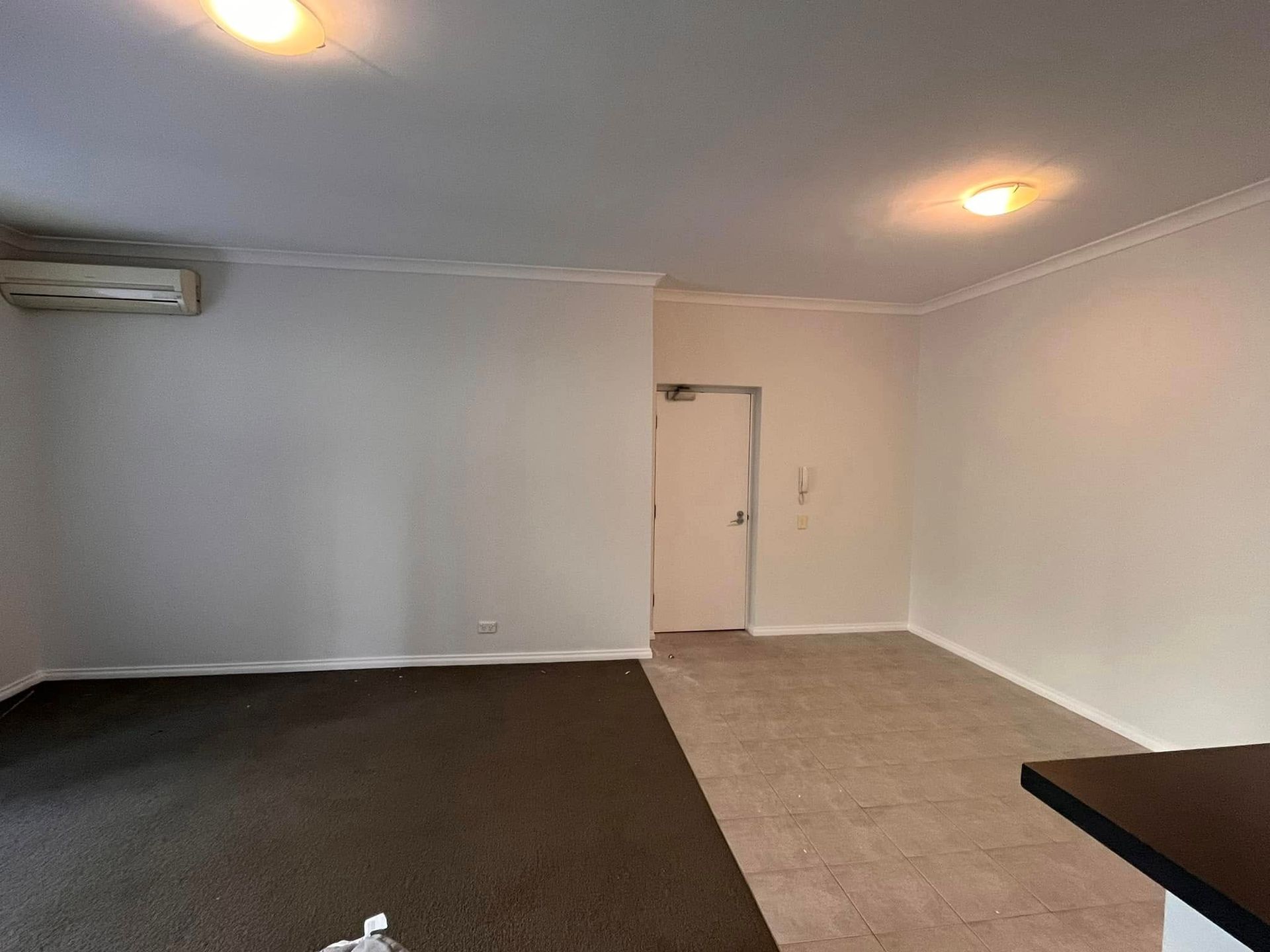 Empty living room with white walls, beige carpet, dark flooring, ceiling lights, and a wall-mounted air conditioner.