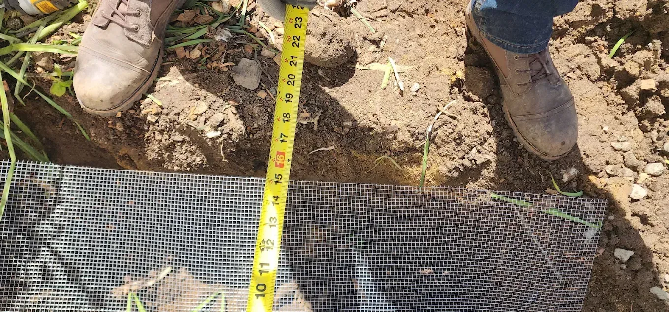 Boots and measuring tape over a wire mesh laid in a dirt trench during construction work
