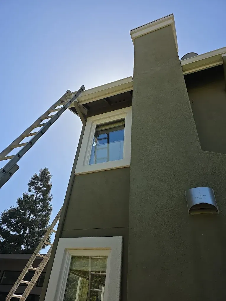 Ladder leaning against an olive-green house beside a tall chimney and windows