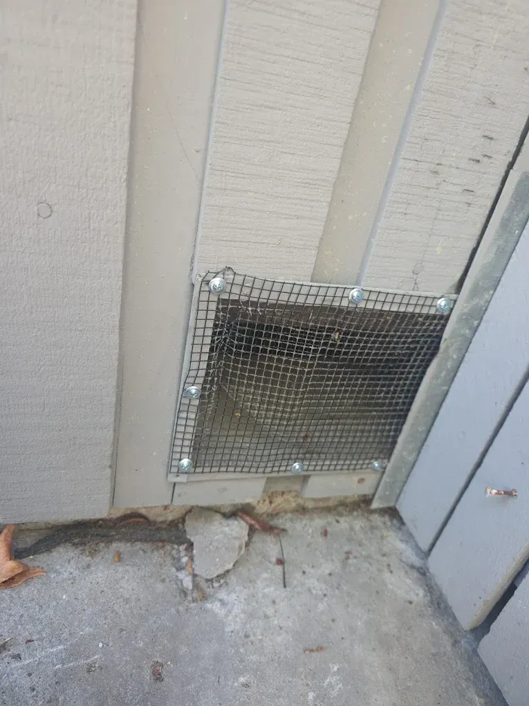 Broken vent cover at the base of an exterior wall with debris on the ground.