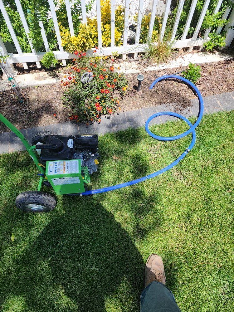 Green pressure washer with blue hose on grass by flower bed and white fence